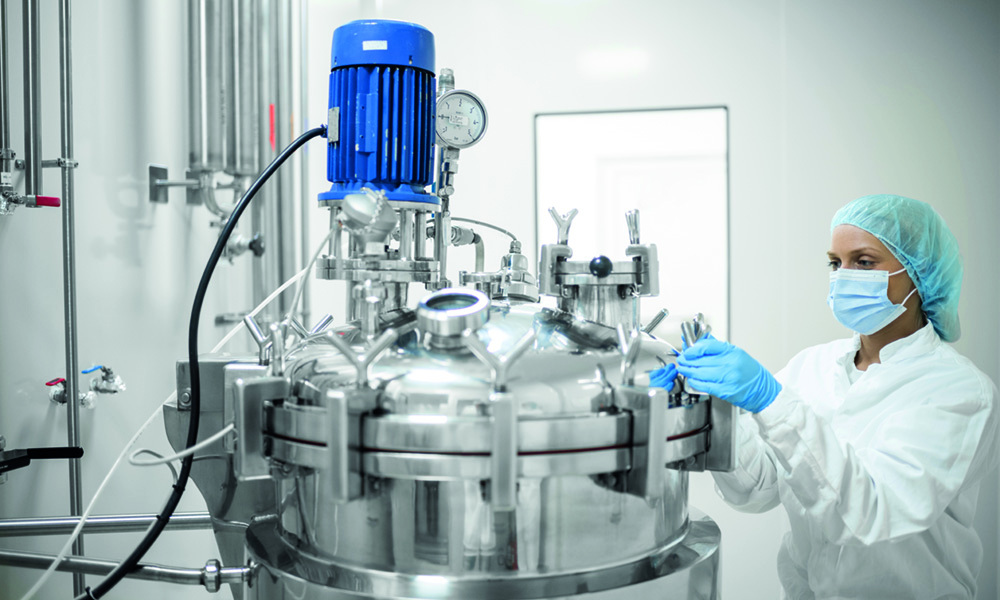 A young female skilled worker in protective gear operates a high-tech equipment in a pharmaceutical production facility, ensuring the precise mixing of substances to produce life-saving medications. Highlighting the dedication and precision required in the industry.