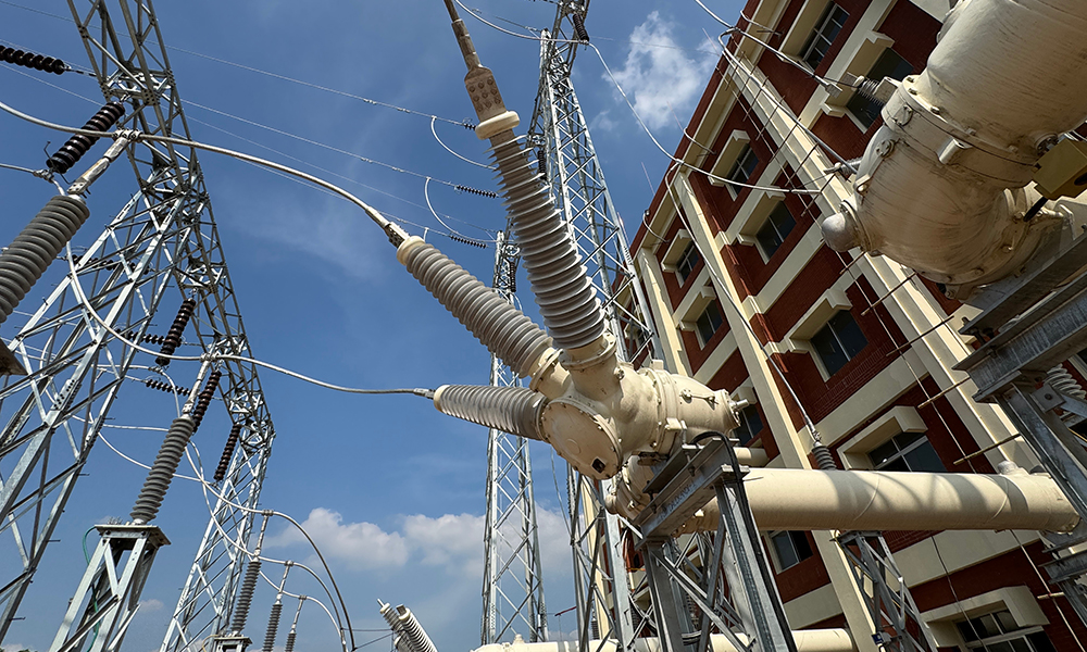 High-voltage electrical substation with insulators, circuit breakers, and steel structures near a multi-story building under clear blue sky.