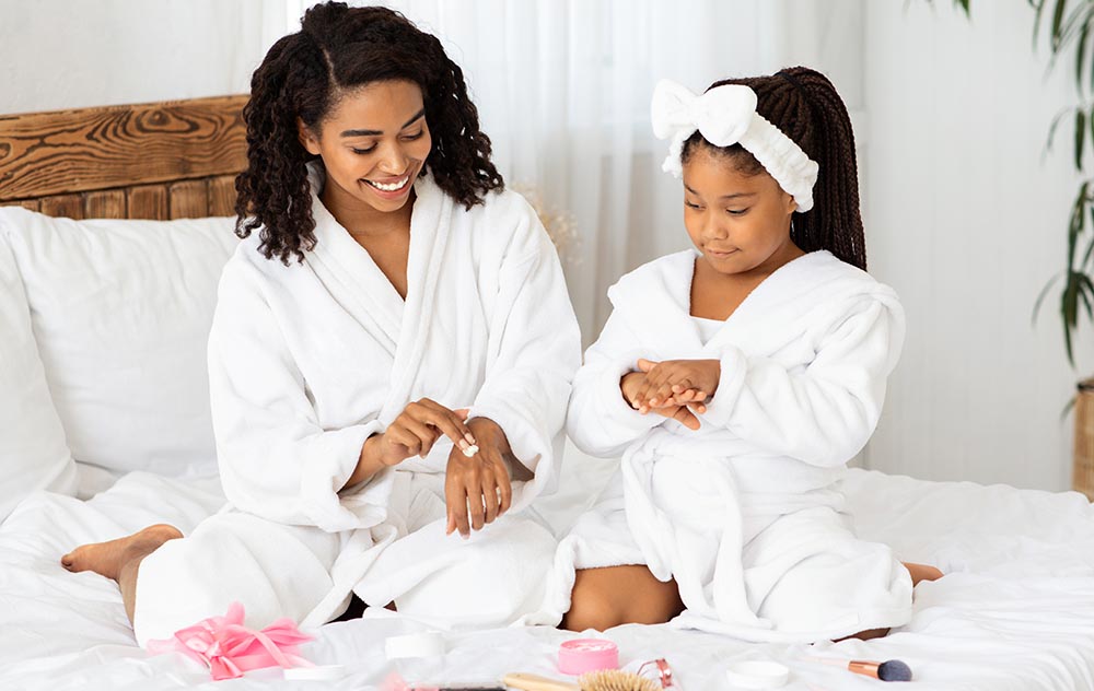 Mother Daughter Beauty Treatments. Caring Black Mom Teaching Little Girl Self-Care At Home, Sitting In Bathrobes On Bed, Applying Body Lotion On Hands, Different Makeup Cosmetics Lying Around